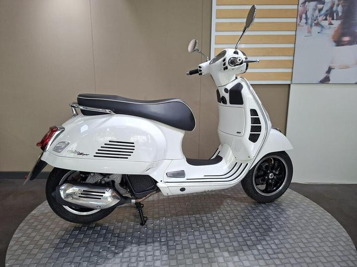 A white Vespa scooter is displayed on a textured circular platform, showcasing its sleek design and black accents against a neutral background.