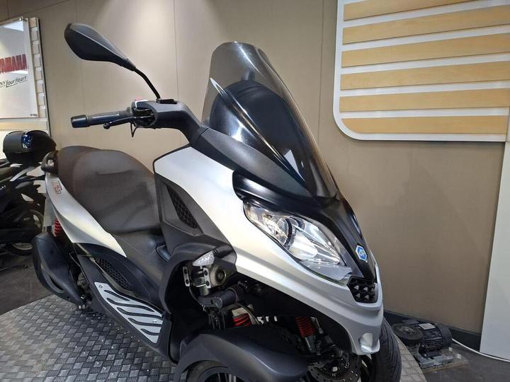 A sleek, silver and black scooter with a large windscreen, parked on a checkered floor in a showroom setting. The handlebars and seat are prominently visible, and other scooters can be seen in the background.