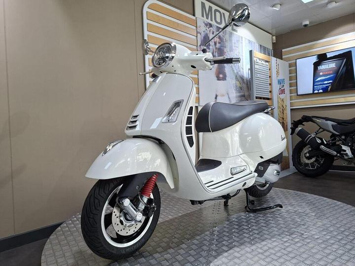 A sleek white scooter is displayed in a showroom, featuring a rounded body, chrome details, and a comfortable seat. The background includes promotional signage and another motorcycle is partially visible to the right.