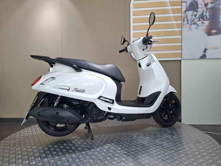 A side view of a white scooter with black accents, featuring a sleek design and modern styling. The scooter is positioned on a textured circular platform with a neutral background.