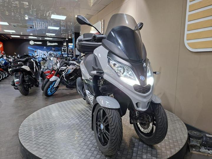 A gray three-wheeled scooter displayed on a circular platform in a motorcycle showroom, surrounded by various other bikes, under bright lights and a sleek interior.