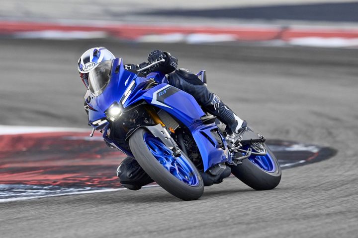 A racer in black gear leans into a turn on a bright blue sport motorcycle, with the track's curbing visible beneath them.