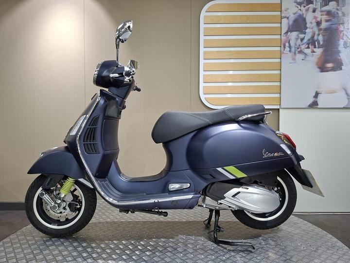 A blue Vespa scooter is displayed side-on in a modern showroom with soft lighting, featuring chrome mirrors, a comfortable black seat, and green accents on the body, set against a wall with a decorative wooden panel and a blurred photograph of pedestrians.