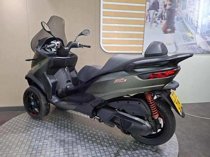 A three-wheeled scooter with a modern design, featuring a dark gray body, a tinted windshield, and sporty accents, positioned on a textured circular display platform. In the background, there is a blurred image of a cityscape with people walking.