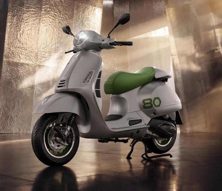 Vespa Edizione Ottantesimo A modern scooter with a sleek silver body and a green seat, set against a glossy, metallic background, showcasing its stylish design.