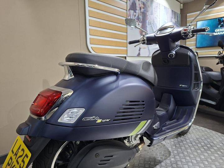 A dark blue scooter, featuring a shiny chrome handlebar and seat, is parked on a textured surface. The rear view displays a prominent tail light and a license plate. The background includes blurred images of urban scenes and additional scooters.