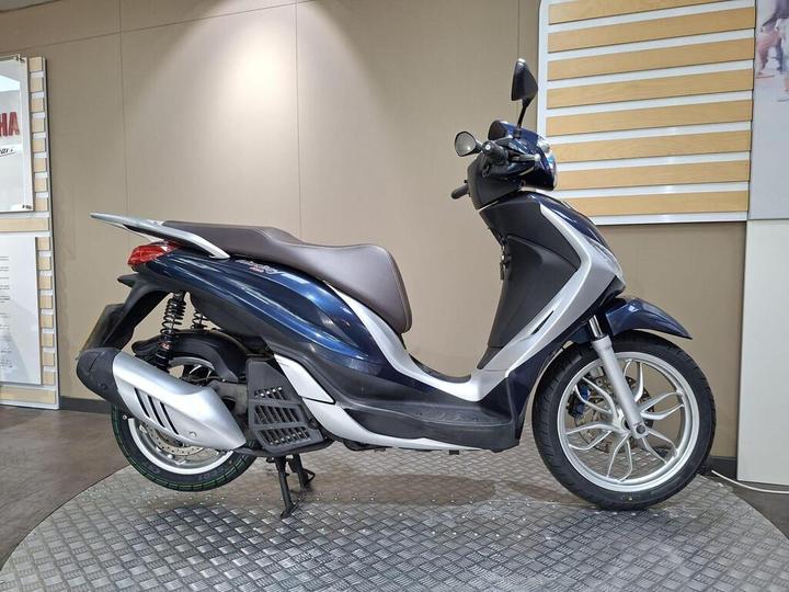 A blue and silver scooter parked on a textured surface, with a simple background featuring light-colored walls and promotional displays.