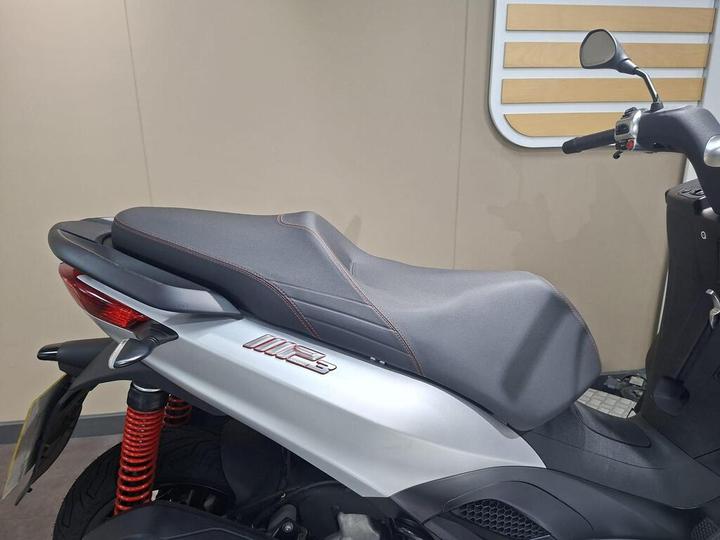 A close-up view of the seat and rear of a modern scooter, featuring a sleek design with a gray seat and red accents, set against a neutral background.