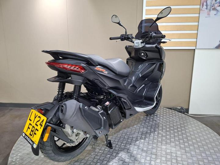 A sleek black scooter displayed at an angle, highlighting its rear and side features, including a distinctive tail light and exhaust. The scooter sits on a textured surface in a neutral-toned setting.