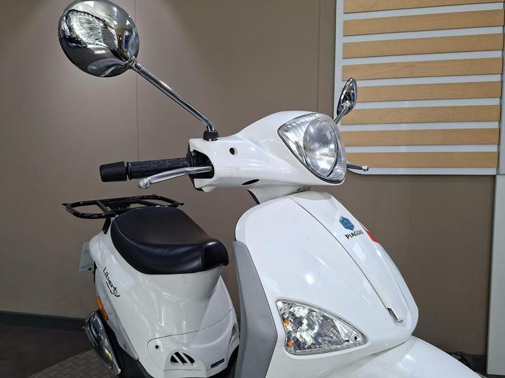 A close-up of a white scooter with a gleaming chrome mirror and headlight, featuring a black seat and the Piaggio logo prominently displayed on the front. The background is neutral and soft, emphasizing the scooter's design.