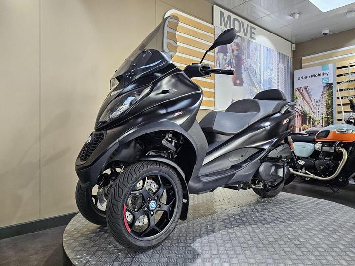 A sleek black three-wheeled scooter displayed on a circular, patterned platform in a modern showroom, with a partially visible motorcycle in the background.