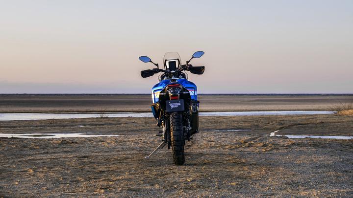 A blue motorcycle is parked on sandy terrain with a river in the background, under a soft sunset sky.