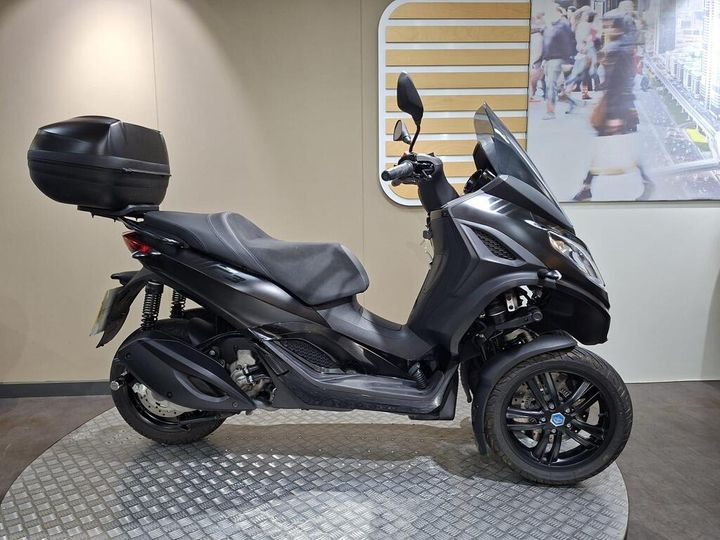Piaggio MP3 300 HPE (2024) A sleek black three-wheeled scooter displayed on a textured platform, featuring a top box for storage and a windshield for protection. The background includes a blurred image of people walking.