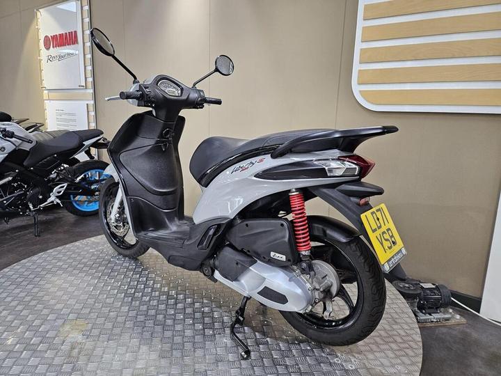 A Yamaha Neo's scooter displayed in a showroom, featuring a sleek gray body, red suspension spring, and a unique license plate; other motorcycles are visible in the background on a checkered floor.