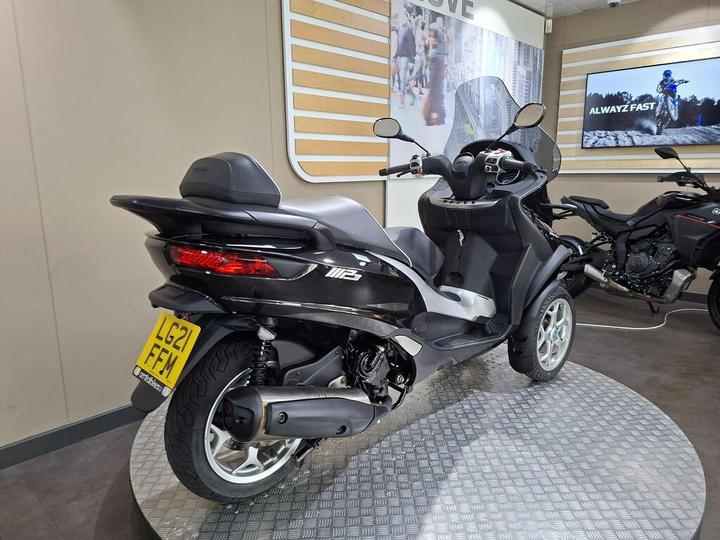 A black scooter with a windshield, parked on a round metal platform, showcasing its rear. The scooter has a license plate visible with the registration number LG21 FFM, and a light gray wall behind it displaying promotional images of motorcycles.