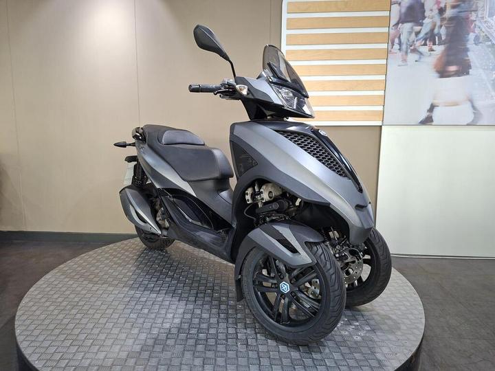 A sleek, gray three-wheeled scooter displayed on a circular metal platform against a neutral wall. The scooter features a modern design with a prominent front shield and stylish wheels.