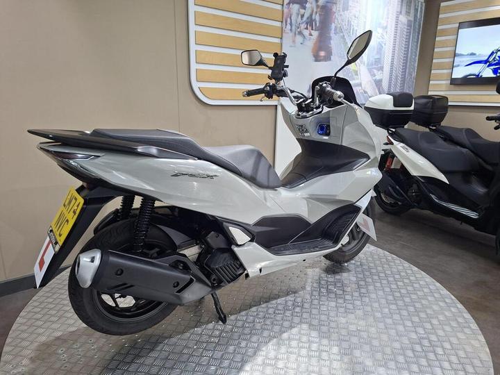 A sleek white scooter displayed on a circular metallic platform, featuring a sporty design with a black seat and modern styling, set against a light-colored wall with promotional images in the background.