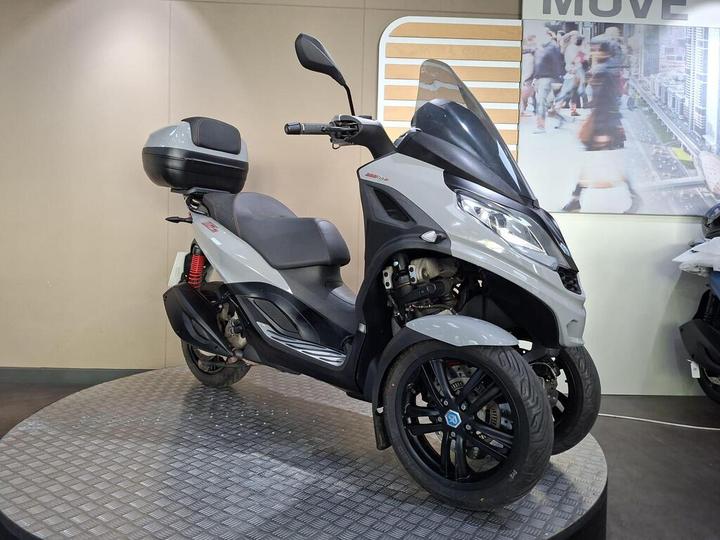 A gray three-wheeled scooter on a round display stand, featuring a large rear storage compartment and a windshield, set against a modern shop interior.