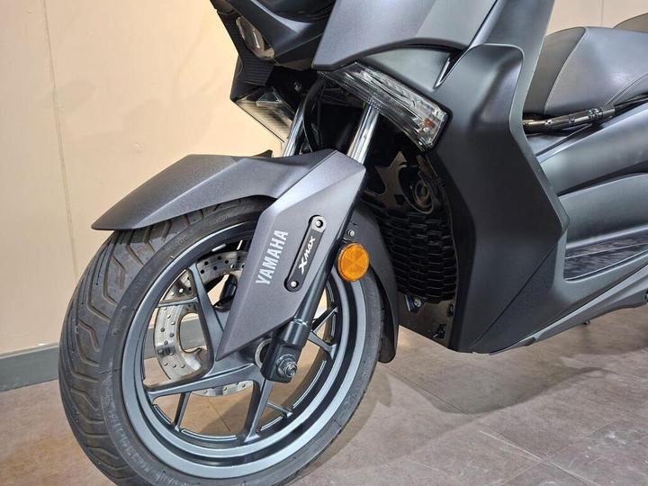 Close-up of the front wheel and fender of a Yamaha Xmax scooter, showcasing the branded hubcap, tire tread, and brake system against a neutral background.
