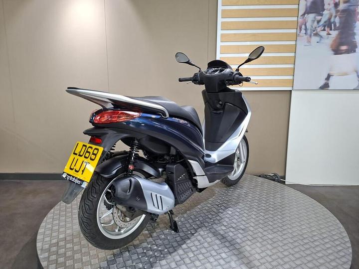 A blue and silver scooter displayed on a circular stand, showing its rear view, with a visible license plate number LD68 WJT. The background features a light-colored wall and a panel with wooden stripes.