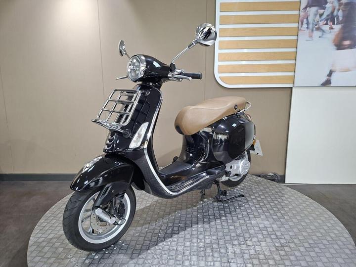 A sleek black scooter with a curved design and tan seat, displayed on a circular metal base, against a neutral background.