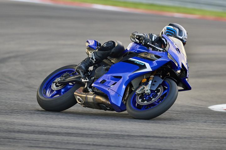 A motorcyclist leans into a turn on a blue sport motorcycle, wearing a black racing suit and helmet, on a racing track.