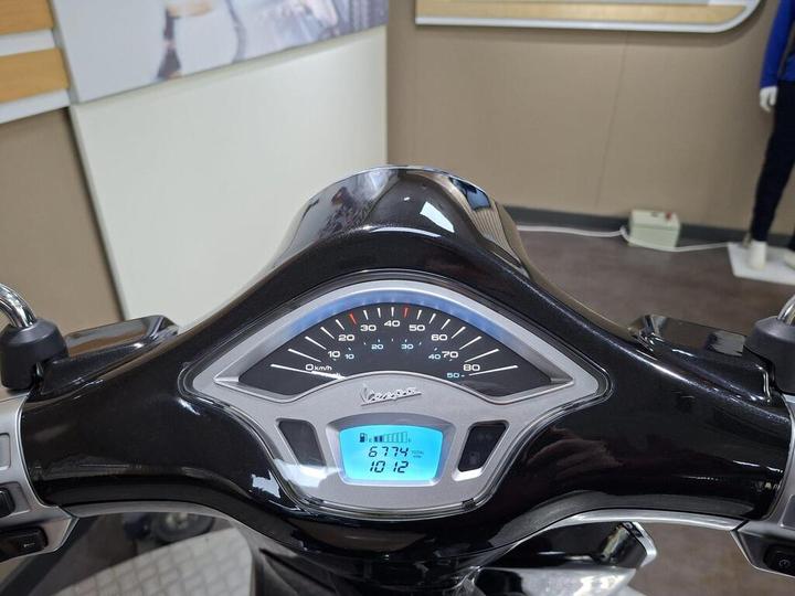 Close-up of a Vespa scooter dashboard displaying speedometer, odometer reading 6,774 km, and a total trip meter showing 10.12 km, set against a blurred backdrop of a show room.