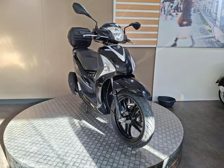 A sleek black scooter displayed on a circular platform in a showroom, with a light background and a partially visible wall poster in the back.