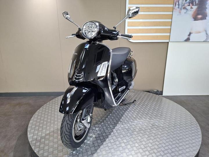 A sleek, black scooter displayed on a rotating circular platform with a textured metal surface, featuring a prominent headlight and chrome details. The background is a plain wall with a subtle design.