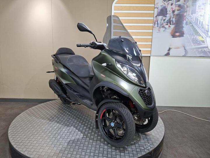 A green three-wheeled scooter with a sleek design is positioned on a circular platform with a metallic surface. The scooter features a windshield, sporty black wheels, and a comfortable seat. In the background, there is a display of people walking in an urban setting.