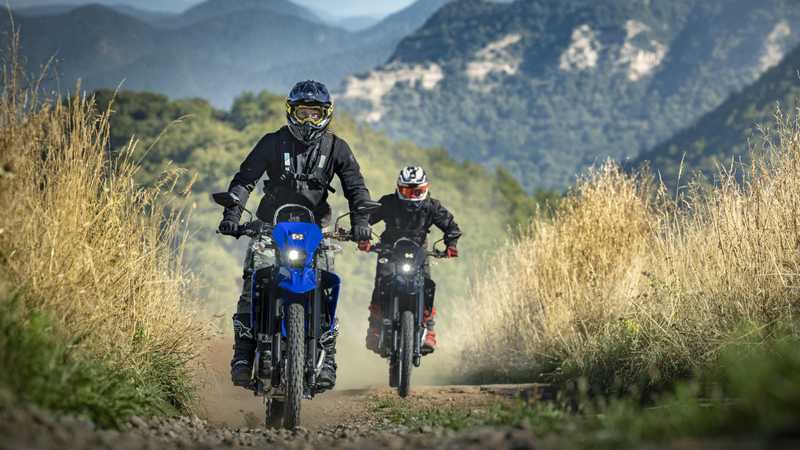 Two motorcyclists ride off-road on dirt trails, with mountains in the background and tall grass lining the path. The lead rider is on a blue motorcycle, while the second rider is on a black motorcycle, both with helmets and protective gear. Dust is kicked up behind them as they navigate the terrain.