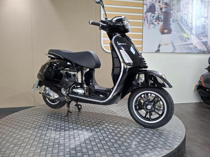 A sleek black Vespa scooter displayed on a circular stand, featuring a glossy finish, modern design, and chrome accents, against a minimalist background.