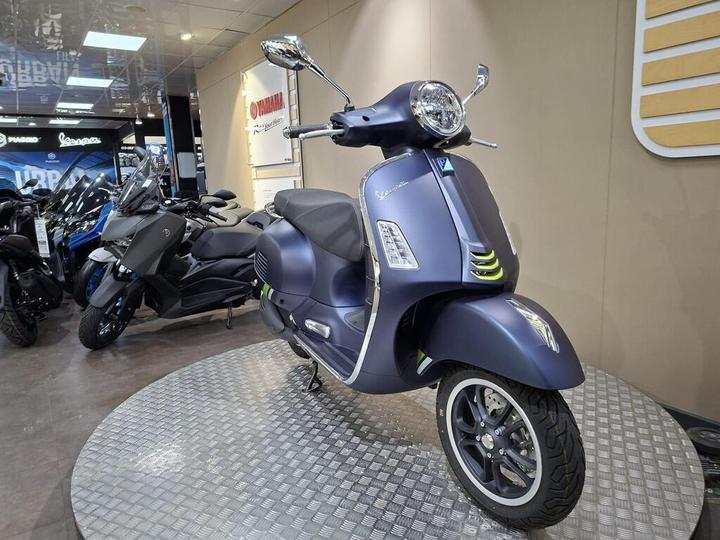 A stylish blue Vespa scooter displayed on a rotating platform in a motorcycle showroom, with various other bikes visible in the background.