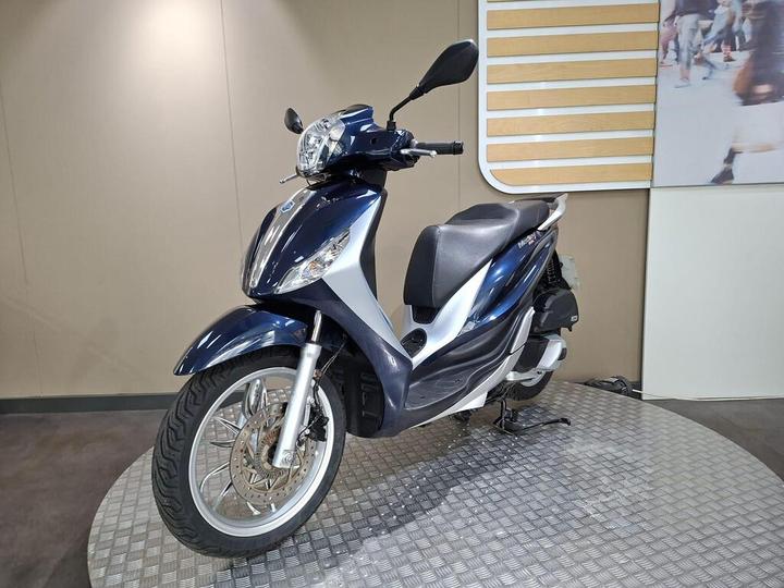 A navy blue scooter is displayed on a circular platform with a textured surface, showcasing its front view, featuring a sleek design and a prominent headlight. The background is neutral, with a subtle wall design.