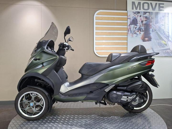 A green three-wheeled scooter displayed in a showroom with a modern backdrop, featuring a transparent windshield and dual seating.