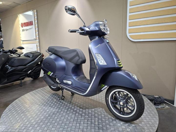 A blue Vespa scooter stands on a circular display, showcasing its sleek design and chrome accents, with a black motorcycle partially visible in the background.
