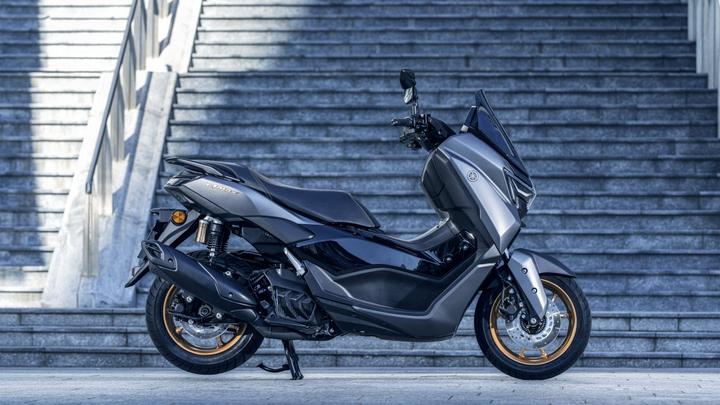 A sleek black and gray scooter with gold-accented wheels is parked on a stairway, showcasing its modern design and details like a windshield and stylish body lines.