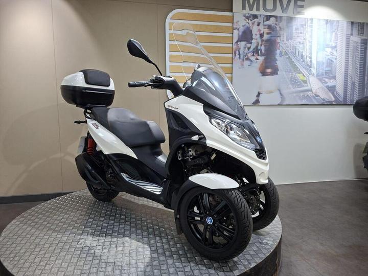 A white and black three-wheeled scooter displayed on a circular platform, with a transparent windscreen and a top box mounted on the rear, against a backdrop featuring a cityscape.