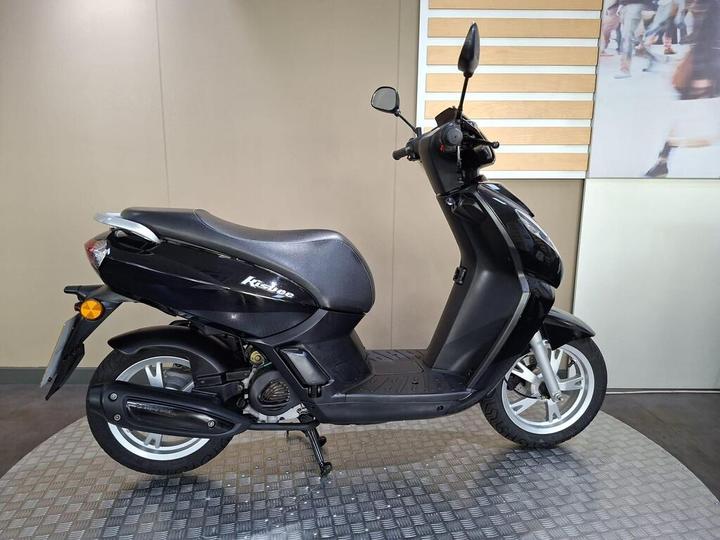 A black scooter is showcased from the side, featuring a sleek design and a comfortable seat, positioned on a textured surface with a neutral background.