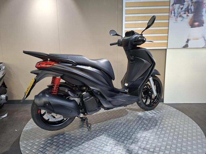 A sleek black scooter is displayed on a circular diamond-plate surface, showcasing its modern design and red suspension springs. Soft lighting highlights its contours against a beige backdrop with a partially visible wall image.