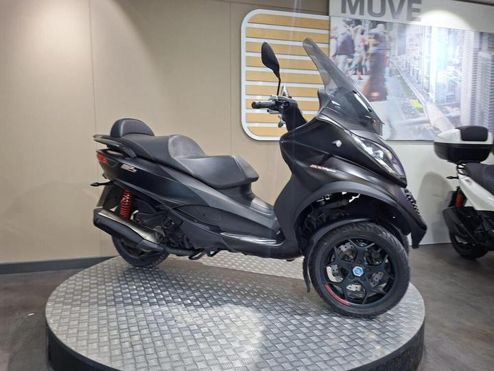 A sleek black scooter with a modern design is displayed on a circular platform, set against a neutral wall backdrop. The scooter features a windshield, dual seating, and is positioned with one wheel at the front and two at the back.