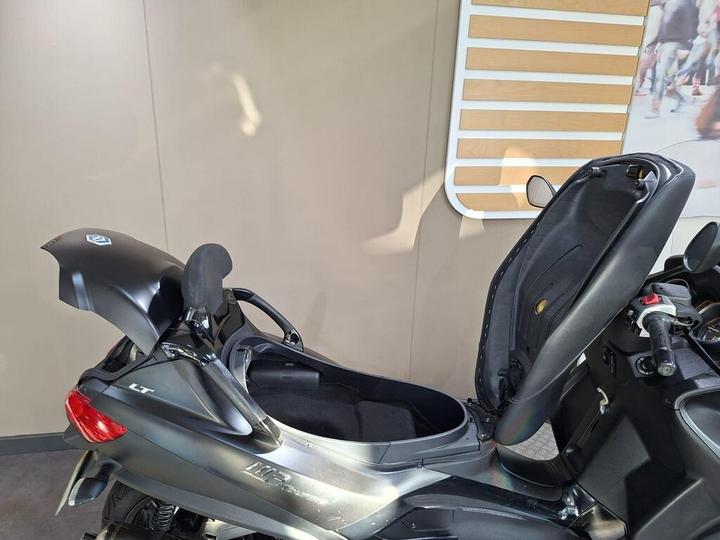 A black motorcycle with its seat raised, revealing a storage compartment underneath, set against a neutral background.