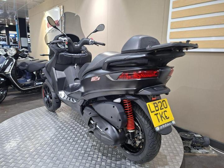 A black scooter with a sleek design is shown from the rear, featuring red suspension springs and a visible license plate reading "LB20 TKE." In the background, another scooter is partially visible, and the setting appears to be a showroom with a textured floor.