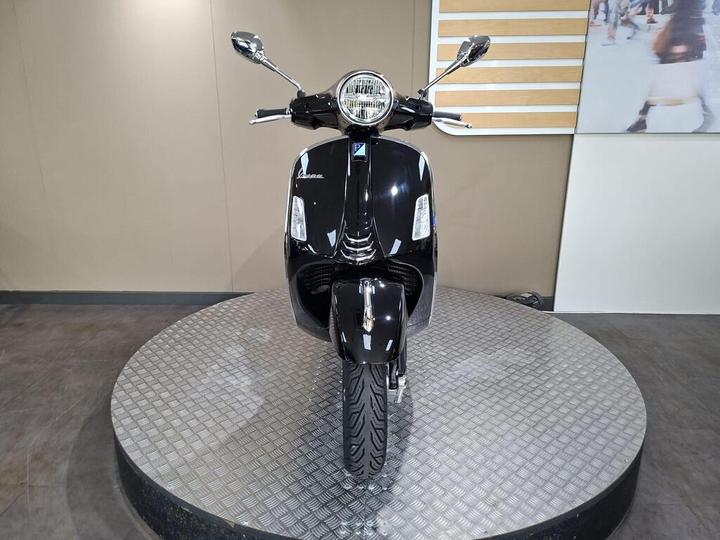 A black scooter is displayed front-facing on a rotating platform, showing its chrome details and front wheel. The background is a neutral color with a small section of a white wall.