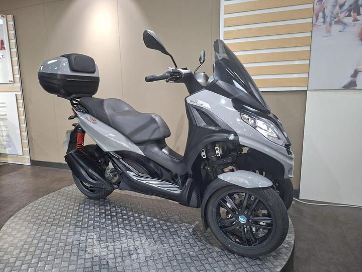 A gray three-wheeled scooter with a sleek design is displayed on a circular platform in an indoor showroom. The scooter features a black storage compartment on the back, a comfortable seat, and modern headlights.