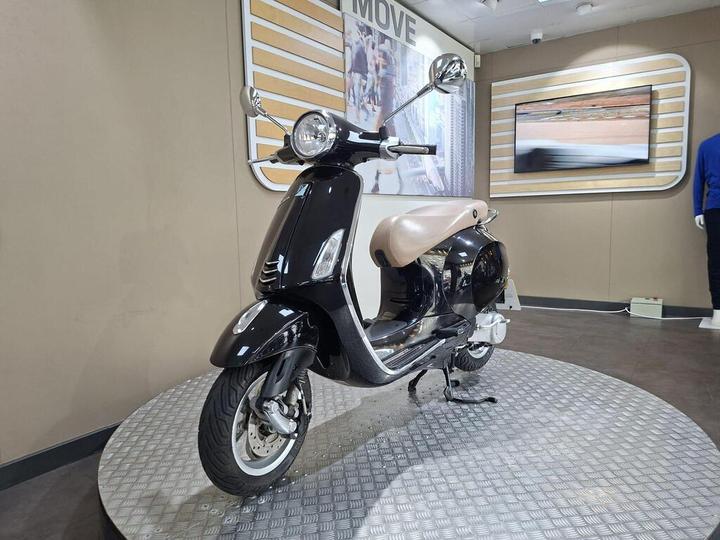 A black and beige scooter displayed on a circular metal platform in an exhibit, with a modern interior featuring a wall-mounted screen in the background.