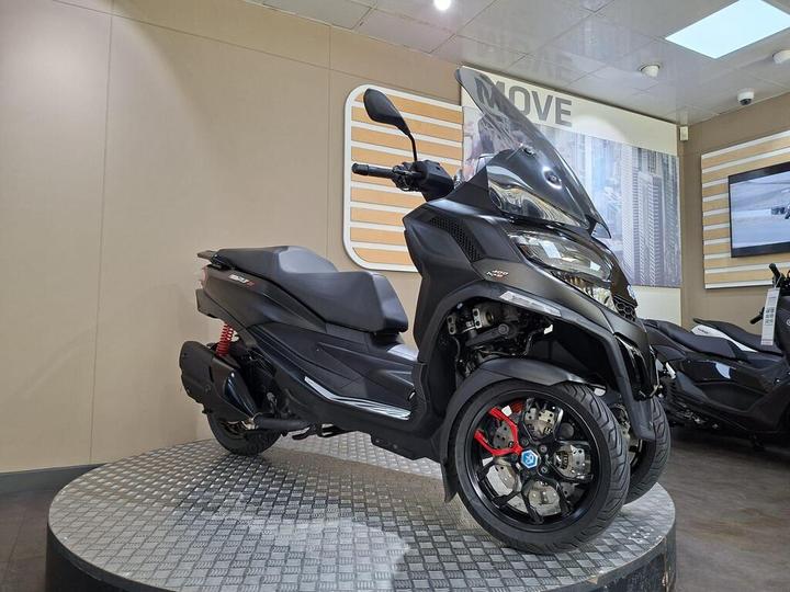 A sleek black scooter with three wheels is displayed on a circular platform inside a showroom. The scooter features modern design elements, including a windshield and prominent red brake calipers. Background displays include wall panels and an additional scooter.