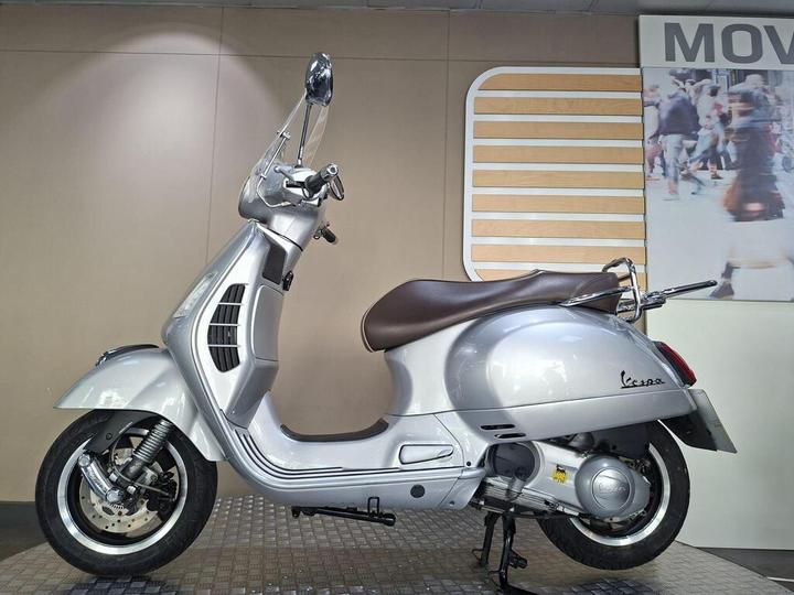A sleek silver Vespa scooter is positioned at an angle against a neutral background, showcasing its elegant design and distinctive features, including a brown seat, chrome details, and a prominent front headlight.