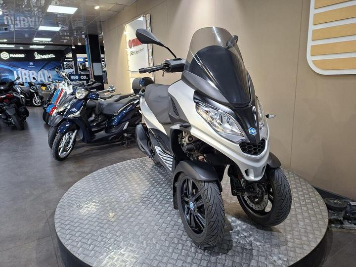 A stylish three-wheeled scooter in silver and black is prominently displayed on a circular platform, surrounded by various other scooters in a well-lit showroom.