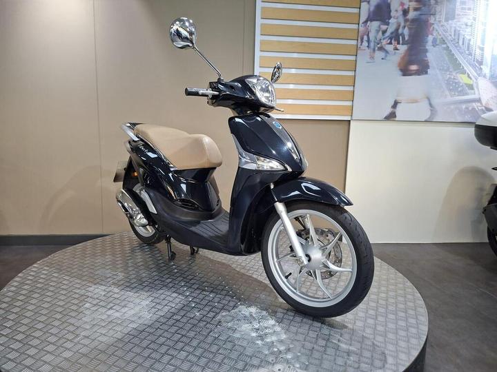 A sleek black scooter with a beige seat, positioned on a textured circular display platform against a neutral background.